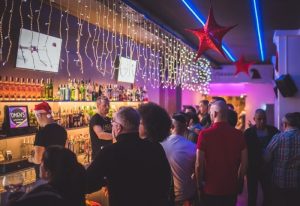 Dmen's Gay Bar in Barcelona: Festive interior view with a crowd of gay men, barman, Christmas lights, and holiday decorations. Gay travel advice.