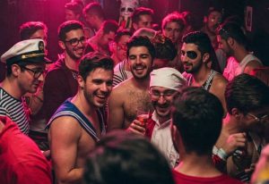 Crowd at Moeem Barcelona, a gay bar in Barcelona. Men in sailor costumes and casual wear smile and socialize under pink lighting.