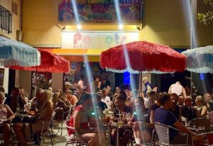 Parrots Pub terrace, a popular gay bar in Sitges, Spain. People sit at tables under colorful umbrellas. Nightlife scene.