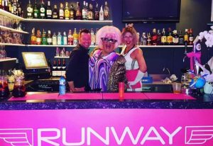 Runway Terrace Bar, a gay bar in Sitges. Staff and drag queens behind the bar with the Runway logo. Gay travel advice.