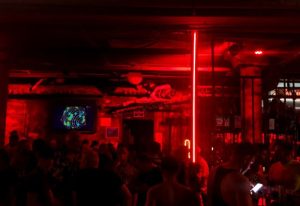 Inside Strong Construction gay cruise club in Gran Canaria. Red lighting, crowd of men, screen, bar. Gay nightlife travel.