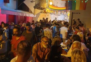Busy gay bar in Ibiza at night, rainbow flags overhead. Gay men and women socialize at tables. Sunrise Ibiza gay bar scene.