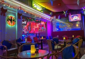 Interior of Zaza Bar, a gay bar in Gran Canaria. Tables, chairs, festive decor, and a Bacardi logo are visible. Nightlife scene.