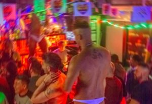 Afterline Milan: Gay men dancing in a crowded, colorful club. Nightlife scene with shirtless men and rainbow decorations. Gay travel advice.