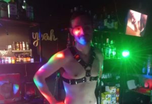 Drako Club Lisbon: Man in harness at gay bar, with colorful lights and bar shelves in background. Gay travel advice for Lisbon nightlife.