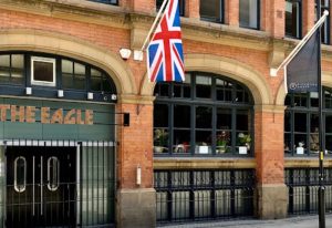 The Eagle Manchester, a gay bar, with a British flag. Brick building, arched windows, and dark trim. Gay travel advice.