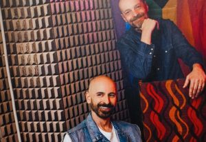 Mono Bar Milan portrait. Two smiling men, one in a denim vest, the other in a shirt, pose in front of a geometric wall. Gay travel.