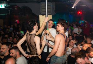ONEWAY Discoclub Milan: Gay men dancing at a crowded party. Some are shirtless, others wear mesh. A pole is the focus of attention.