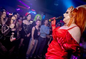 Drag queen performs at On Bar, Manchester. Crowd enjoys the show in the vibrant gay nightlife scene. Red dress, microphone, and stage lights.