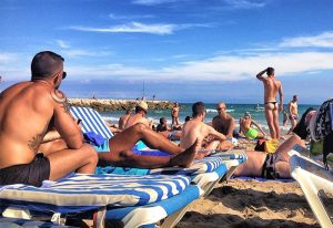 Gay nude beach scene in Sitges. Men sunbathing on a sandy beach, some on loungers, under a blue sky. Popular gay beach destination.