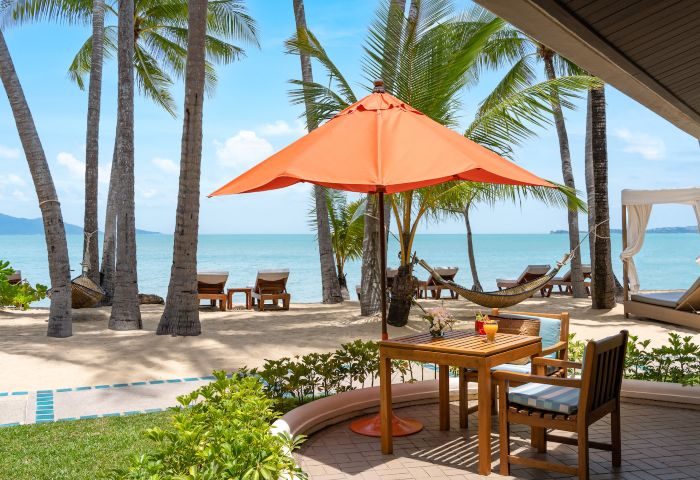 Deluxe Beachfront Villa patio at Santiburi Koh Samui. Plunge pool view, palm trees, beach chairs, orange umbrella, table with drinks, hammock.