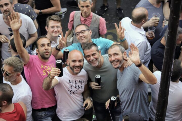 COX: Gay men at a street party, smiling and waving. Group of friends enjoying drinks, celebrating gay pride. Travel advice for gay men.