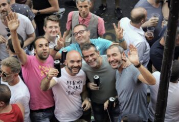 COX: Gay men at a street party, smiling and waving. Group of friends enjoying drinks, celebrating gay pride. Travel advice for gay men.