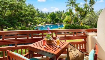 Santiburi Koh Samui two-bedroom duplex suite balcony view. Drinks, table, chairs, and a bag overlook the pool and lush tropical landscape.