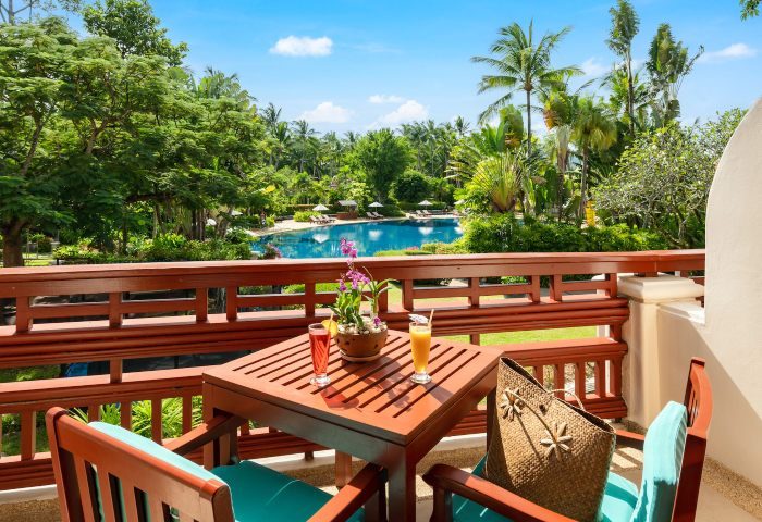 Santiburi Koh Samui two-bedroom duplex suite balcony view. Drinks, table, chairs, and a bag overlook the pool and lush tropical landscape.