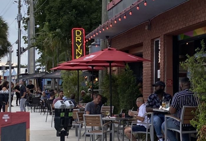 Outdoor seating area at The PUB Wilton Manors. Patrons enjoy drinks under red umbrellas. A neon DRYNK sign glows above. Gay travel.