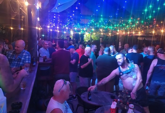 Gay men at Scandals Saloon, a lively bar. Rainbow lights adorn the ceiling. A man in a harness holds a drink. Travel advice. 1-3.jpg