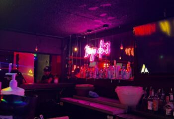 Inside 11:11 Club, a gay bar in Cancun. Neon signs, bottles on the bar, and a bartender create a vibrant, inviting atmosphere.