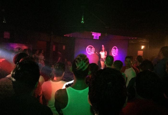Inside 11:11 Club, a gay bar in Cancun. A performer on stage entertains a crowd of gay men. The scene is dark with colorful lighting.