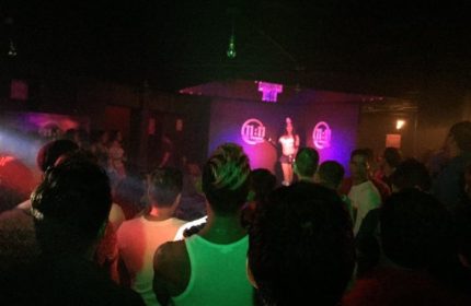 Inside 11:11 Club, a gay bar in Cancun. A performer on stage entertains a crowd of gay men. The scene is dark with colorful lighting.