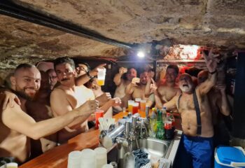 Gay men at Bears' Den bar. A group of shirtless men, some with beers, pose behind a bar in 14_bearsden_02. Gay travel advice.