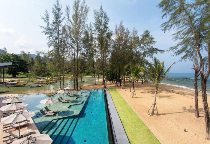 La Vela Khao Lak resort view. Poolside relaxation on beach, lush trees, and boats on river. Gay travel advice for Thailand vacation.