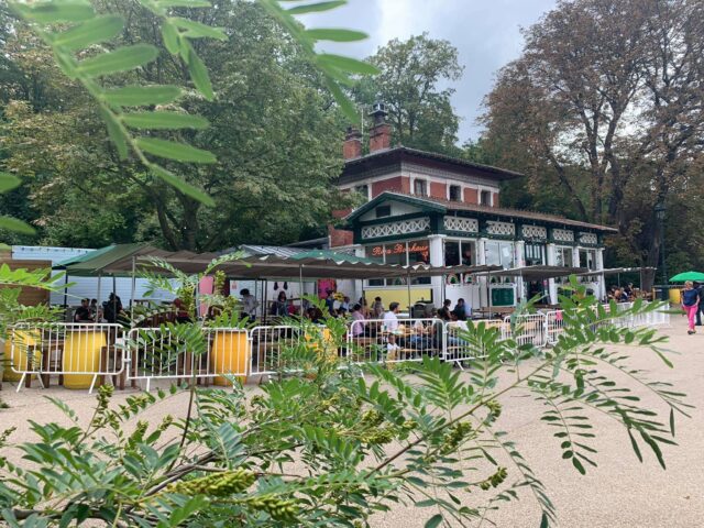 Rosa Bonheur: Outdoor Parisian cafe scene. People at tables, greenery, and architecture. Popular gay-friendly spot for drinks and socializing.