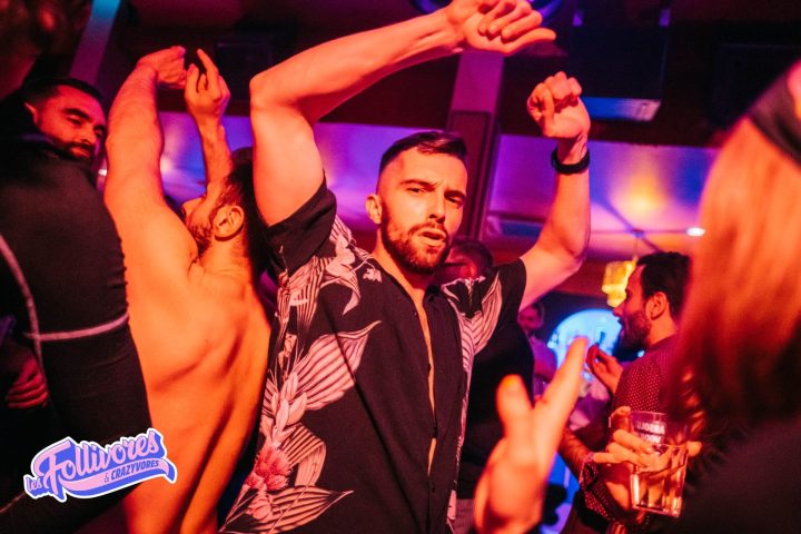 Gay men dancing at La Nuit des Follivores/Crazyvores party. Nightlife scene with shirtless man, club lights, and festive atmosphere. Lesfollivores.