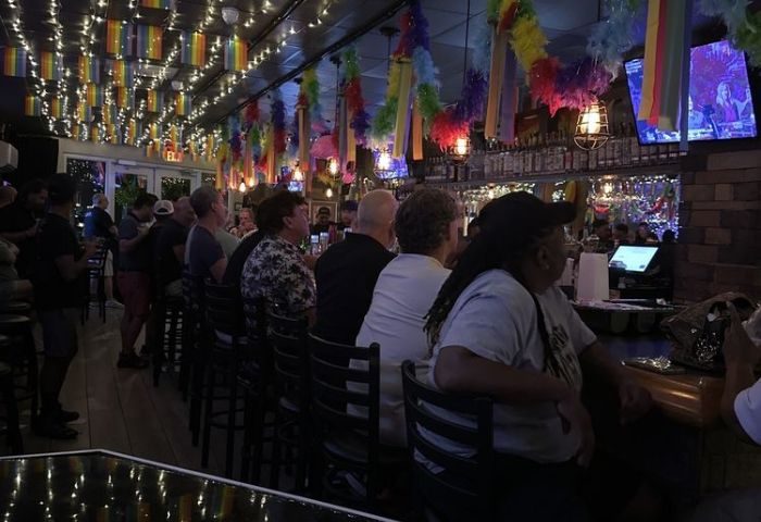 Interior view of The PUB Wilton Manors, a gay bar. People sit at the bar under rainbow flags and colorful decorations. 3-1.jpg.