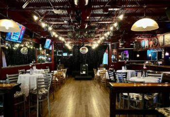 Interior of Scandals Saloon, a gay bar. Tables with white cloths, wooden chairs, stage, and bar area. String lights add to the ambiance.