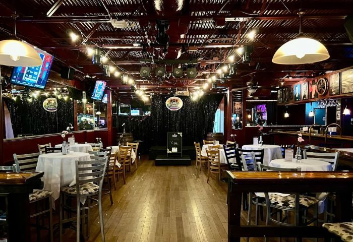Interior of Scandals Saloon, a gay bar. Tables with white cloths, wooden chairs, stage, and bar area. String lights add to the ambiance.
