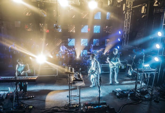 Live band performing in New York. Musicians playing keyboards, guitar, and drums on stage with bright lights and a digital backdrop.