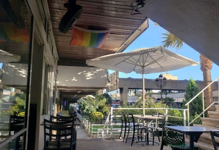 Outdoor seating at 3 Monkeys gay bar in Torremolinos. Rainbow flag, tables, chairs, and parasol visible. Gay travel destination.