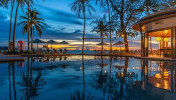 SAii Laguna Phuket pool at dusk. Palm trees, lounge chairs, and a red phone booth line the beach. Reflections shimmer in the water.