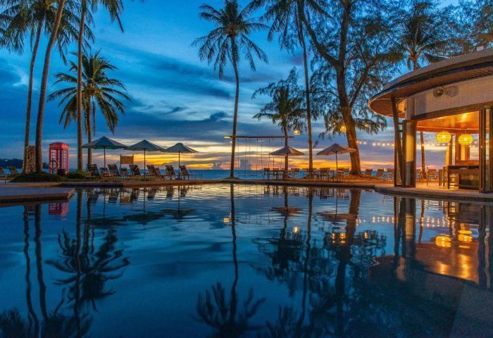 SAii Laguna Phuket pool at dusk. Palm trees, lounge chairs, and a red phone booth line the beach. Reflections shimmer in the water.