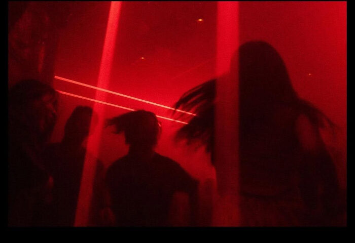 Silhouetted figures in a red-lit club. Nightlife scene for gay men's travel. Potent atmosphere, dance floor, party.