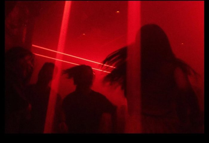 Silhouetted figures in a red-lit club. Nightlife scene for gay men's travel. Potent atmosphere, dance floor, party.