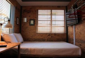Gaythering Sauna room interior. Double bed with white sheets, desk, lamp, and window with blinds. Unique wood paneling. Gay travel.