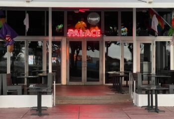 Palace Bar exterior. Neon Palace sign above doors, Halloween decorations. Tables and chairs outside. Gay travel destination.