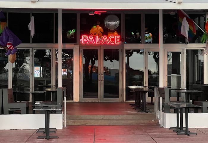 Palace Bar exterior. Neon Palace sign above doors, Halloween decorations. Tables and chairs outside. Gay travel destination.
