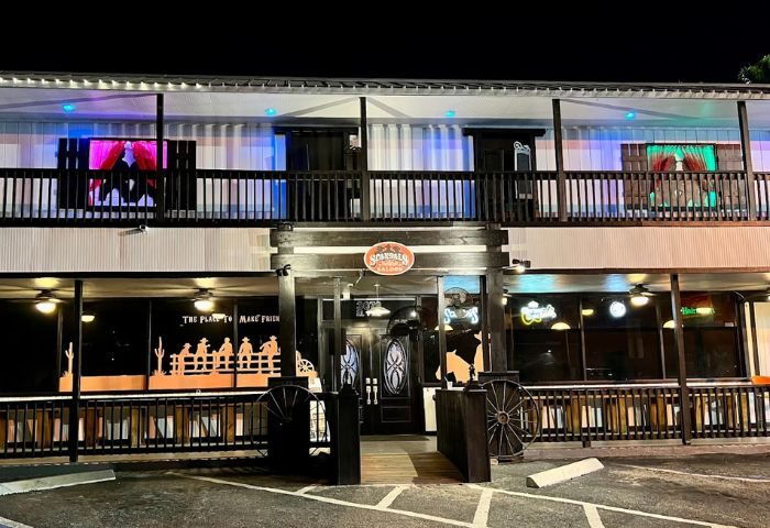Scandals Saloon, a gay bar. Two-story building with Western decor, neon signs, and cowboy silhouettes. Night scene at this gay travel destination.