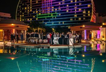Hunters Wilton Manors: Night view of a pool party with people at the bar, colorful lights reflecting in the water, and a building in the background.