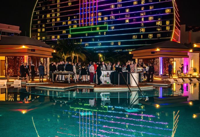 Hunters Wilton Manors: Night view of a pool party with people at the bar, colorful lights reflecting in the water, and a building in the background.