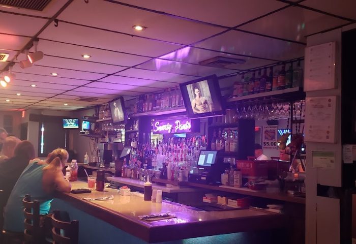 Smarty Pants Bar interior. Gay travel advice: bar with neon sign, TVs showing shirtless men, liquor bottles, and patrons at the counter.