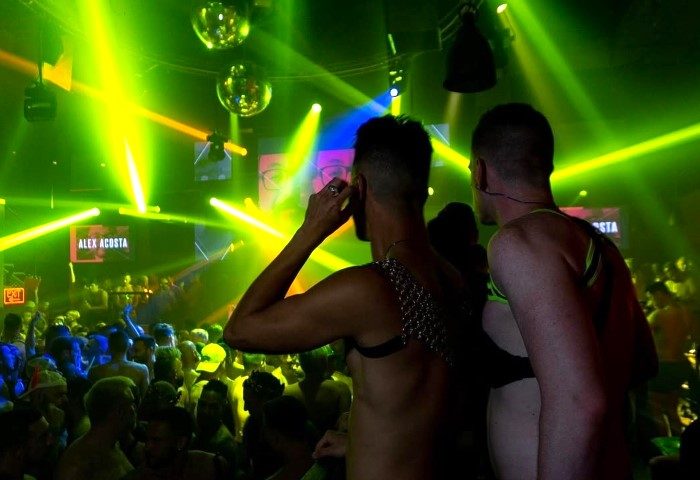 Gay nightlife at Industry, a gay bar. Two men in harnesses face a crowd under green lights and disco balls. Gay travel advice.
