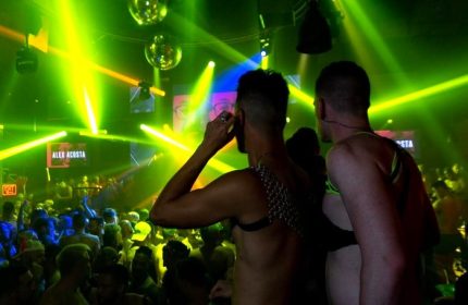 Gay nightlife at Industry, a gay bar. Two men in harnesses face a crowd under green lights and disco balls. Gay travel advice.