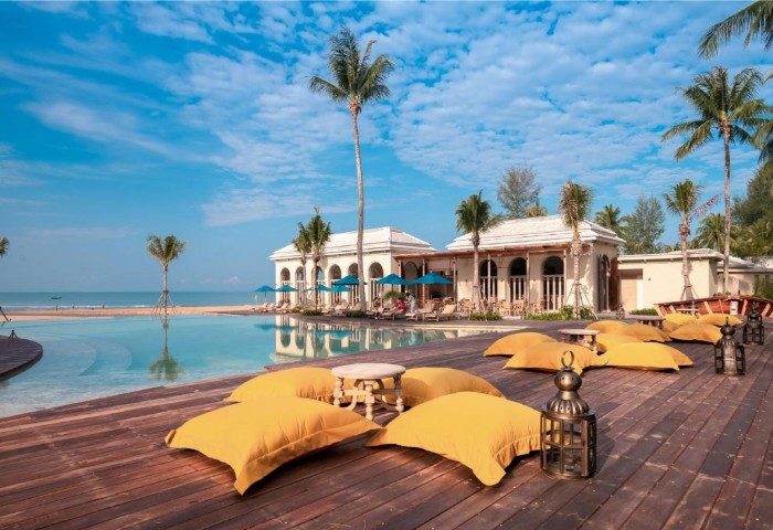 Devasom Khao Lak Beach Resort & Villas pool view. Relaxing yellow pillows, palm trees, and blue sky. Gay travel advice.