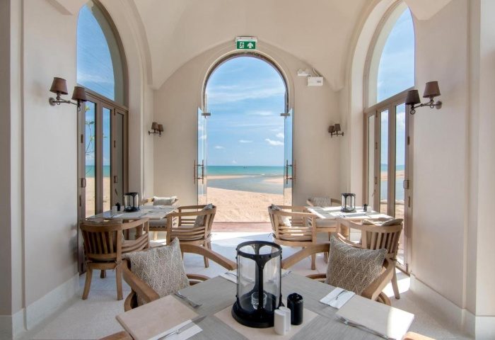 Devasom Khao Lak Beach Resort dining with ocean view. Tables and chairs inside, beach visible through arched doorways. Gay travel advice.