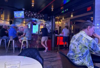 Hotel Gaythering bar scene. Men gather, some at the bar, others seated. Tropical shirts, casual attire. Nightlife, travel.