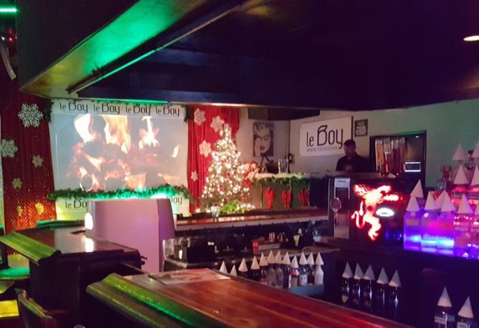 Interior of LeBoy, a gay bar, decorated for Christmas with a tree, lights, and snowflakes. A screen shows a fireplace. Gay travel advice.